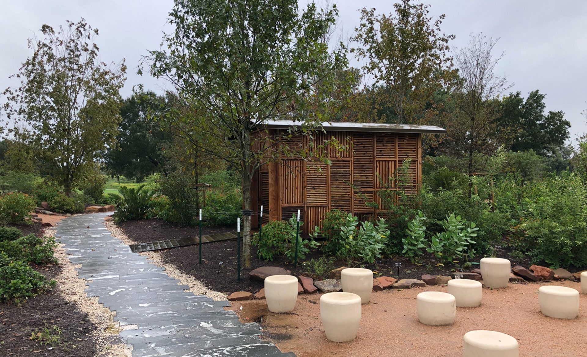 Wooden slat structure in a natural setting at Houston Botanic Garden, featuring white stone stools on a path and greenery in the background Wooden slat structure in a natural setting at Houston Botanic Garden, featuring white stone stools on a path and greenery in the background