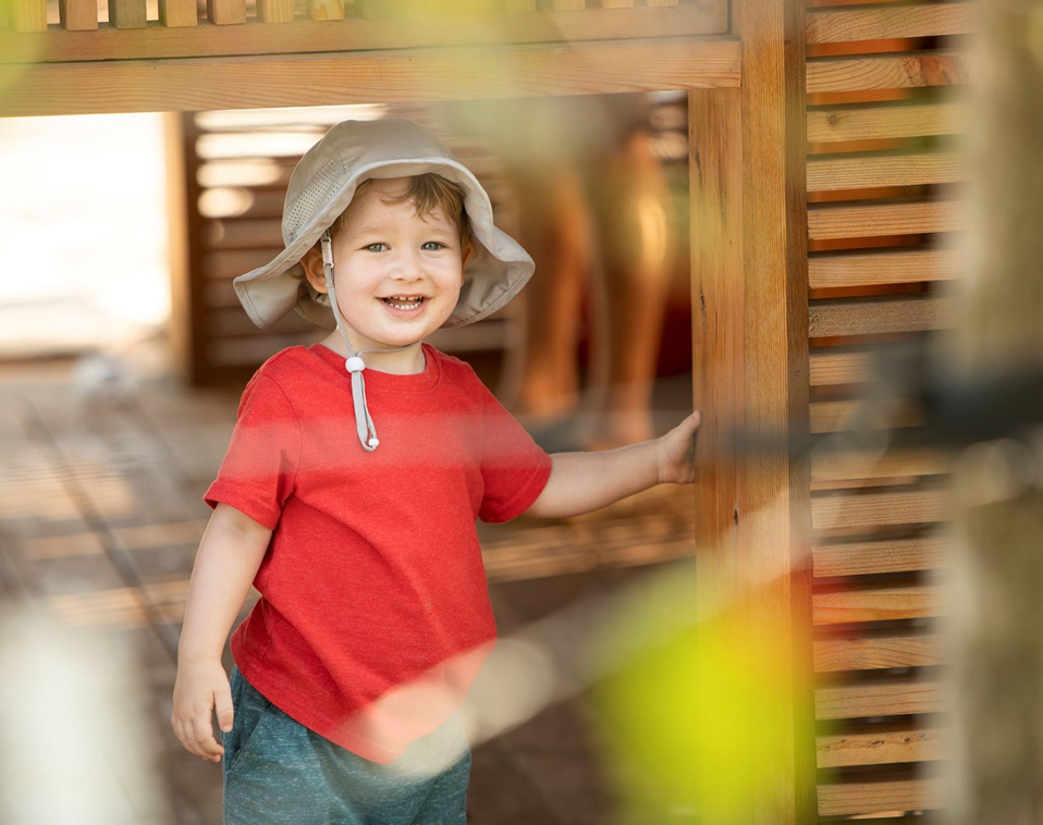 Close-up of a young boy smiling while holding onto a wooden column of a natural structure at the Houston Botanic Garden