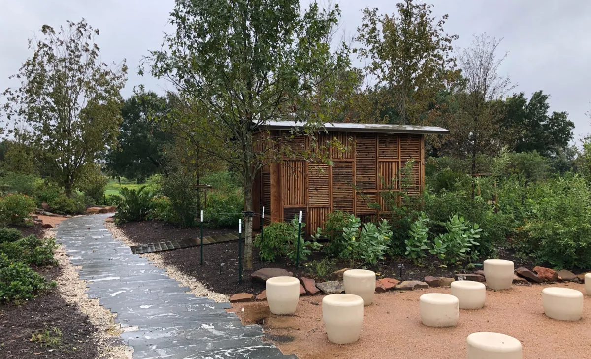Wooden slat structure in a natural setting at Houston Botanic Garden, featuring white stone stools on a path and greenery in the background